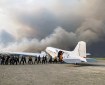 People unload gear from the left side of cargo plane as black and gray wildfire smoke billows in the background.