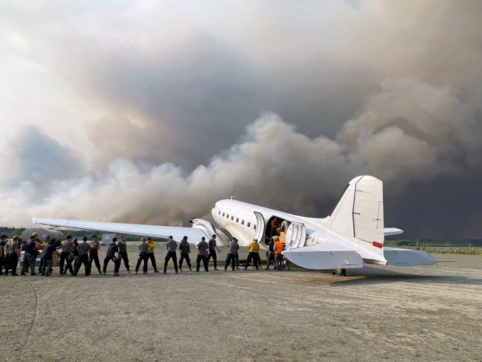 People unload gear from the left side of cargo plane as black and gray wildfire smoke billows in the background.