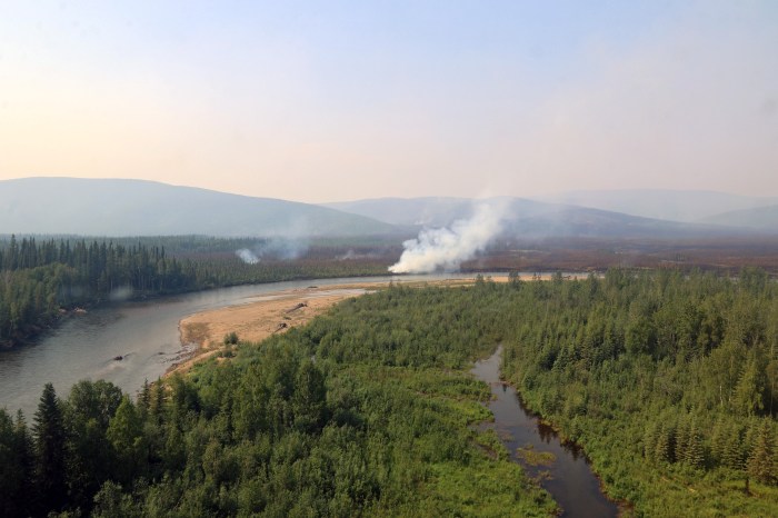 Trees and vegetation are seen in the foreground with a river curving from right to left through the middle of the photo. Wildfire smoke rises into the air from the ground in the middle of the photo.