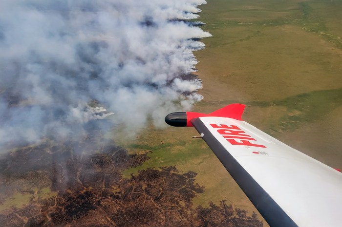 The right wing of a small aircraft with the word FIRE in red on the end of the wing is seen flying above the tundra, some of which is burning with smoke rising into the air.
