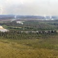 Forest and tundra in the foreground with a river going from left to right through the photo. Wildfire smoke rises from the forested areas in the background.