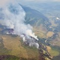 Aerial photo showing wildfire smoke rising from a forested area with a creek on the right side of the image.