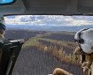 A wildland firefighter wearing a flight helmet sits in the back seat of a helicopter. The helicopter's door is open and the firefighter is using a small, handheld infrared imaging device to scan a burned area for residual heat.