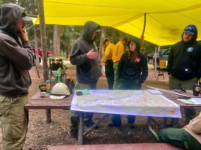 Several people in nomex pants and sweatshirt gather around a table under a yellow tarp, looking at a map.