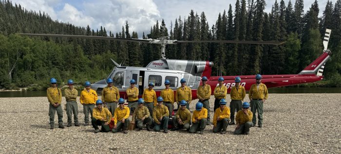 A group of about 20 wildland firefighters poses for the camera in front of a helicopter on a gravel river bed with forest in the background.