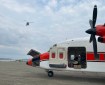 A white plane with a red stripe has landed on an air strip near Rampart to deliver supplies to the Lush Fire. The photo is a close up of the plane, with the nose and prop visible. A helicopter is behind the plane with the line dropped down to the airstrip. Much of the photo is taken up with a sky that is mainly filled with white fluffy clouds. A piece of the Yukon River is visible on the lower left of the photo.