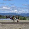 Two firefighters are watering down a helipad to reduce debris for helicopter landing and take off. The helipad is an open gravel area next to the Yukon River. Behind the river are mountains. There is a small helicopter on the pad behind the firefighters. The firefighters are both wearing Nomex protective gear and have a hose stretched out from the river and are wetting the helipad.
