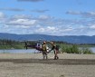 Two firefighters are watering down a helipad to reduce debris for helicopter landing and take off. The helipad is an open gravel area next to the Yukon River. Behind the river are mountains. There is a small helicopter on the pad behind the firefighters. The firefighters are both wearing Nomex protective gear and have a hose stretched out from the river and are wetting the helipad.