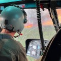 A person in a helicopter is observing fire activity on the Lush Fire. The photo is a close up image in a helicopter. There is a green spruce forest outside with tall orange flames standing straight up and gray plumes of smoke coming up. Inside the helicopter shows the a person observing the fire, they have a jumpsit