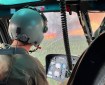 A person in a helicopter is observing fire activity on the Lush Fire. The photo is a close up image in a helicopter. There is a green spruce forest outside with tall orange flames standing straight up and gray plumes of smoke coming up. Inside the helicopter shows the a person observing the fire, they have a jumpsit