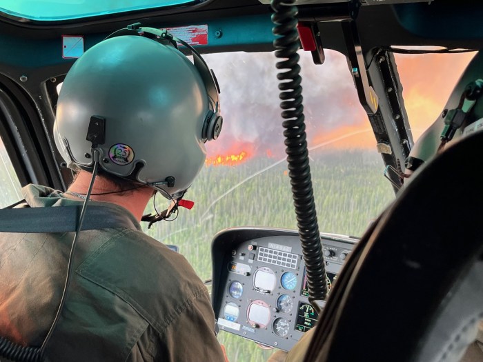 A person in a helicopter is observing fire activity on the Lush Fire. The photo is a close up image in a helicopter. There is a green spruce forest outside with tall orange flames standing straight up and gray plumes of smoke coming up. Inside the helicopter shows the a person observing the fire, they have a jumpsit