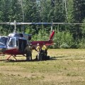 Four wildland firefighters stand outside a helicopter sitting on grass in front of a forested area.