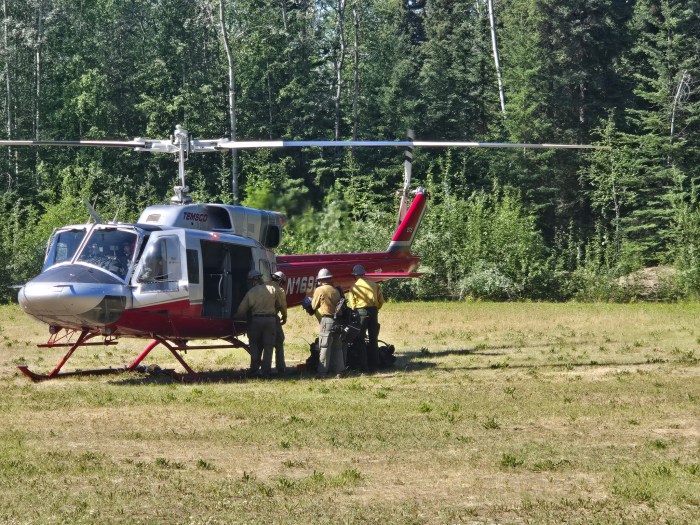 Four wildland firefighters stand outside a helicopter sitting on grass in front of a forested area.