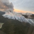 Smoke billows up from a forest fire. Part of an airplane engine is in the photo.
