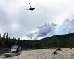 A helicopter with a suspended bucket hovers over a river with a gravel short; a small boat is in the foreground.