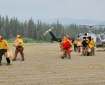 Wildland firefighters wearing yellow shirts and orange hard hats carry gear after unloading gear from a helicopter parked on a gravel area.