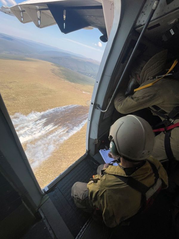 A helmeted person sits in the open doorway of an airplane looking down at smoke billowing up from a grass fire while a person stands beside him, bent over.
