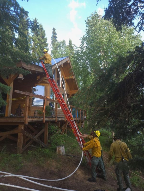 Firefighter on ladder to roof of cabin with hose spraying debris off roof.