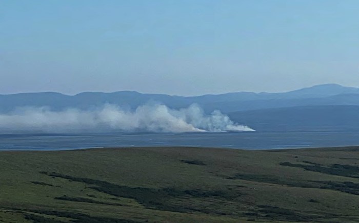 White plumes of wildfire smoke rise from the ground in the distance below rolling hills.
