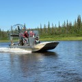 Three people ride in an airboat traveling in a river. A forest of trees is visible on the riverbank behind them.