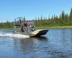 Three people ride in an airboat traveling in a river. A forest of trees is visible on the riverbank behind them.