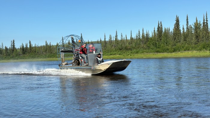 Three people ride in an airboat traveling in a river. A forest of trees is visible on the riverbank behind them.