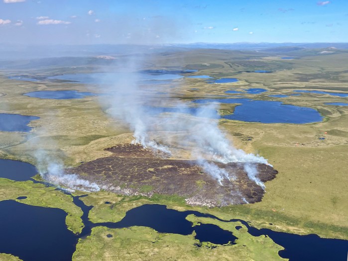 An aerial photo shows wildfire smoke rising into the air from a burned area surrounded by bodies of water.