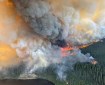 An aerial view showing Birch Creek in the bottom left corner. Flames and a large column of black, gray and white smoke rise from a forested area above the creek.
