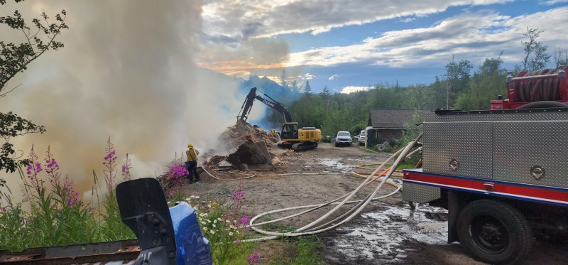 a fire truck with hoses and an excavator work on a smoking pile of debris