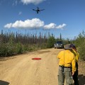 A red circle landing pad is in the center of a dirt road. A drone takes flight, 20 feet in the air. One firefighter uses the control pad, while another firefighter looks over their shoulder. A truck blocks the road in the background.