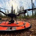 A close up shot of a drone on it's landing pad. The pad sits on wood litter, and there are trees in the background. The sky has scattered clouds and is mostly clear.