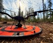 A close up shot of a drone on it's landing pad. The pad sits on wood litter, and there are trees in the background. The sky has scattered clouds and is mostly clear.