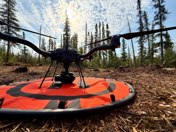 A close up shot of a drone on it's landing pad. The pad sits on wood litter, and there are trees in the background. The sky has scattered clouds and is mostly clear.