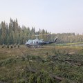 A helicopter, with it's tail to the left side of the image, sits on the ground in a clearing, with a thick stand of trees in the background. A body of water is near the rear of the helicopter. 7 firefighters walk towards the nose of the helicopter, preparing to load.