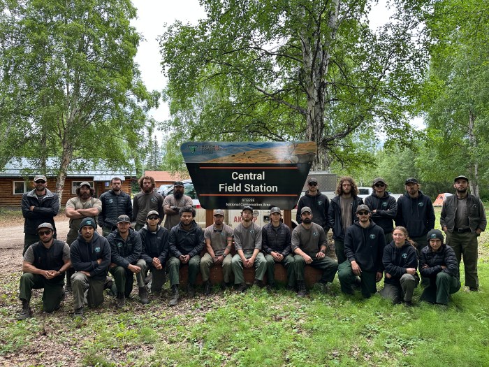 Twenty-two Smith River Hotshots stand and kneel around the Central Field Station sign. Trees and buildings are in the background and they are standing on grass.