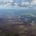 An aerial view of a landscape effected by fire. A braided river, Birch Creek, flows through the center of the image, to the right of the burnt area. A small plume of smoke rises from an area on the fire side of the creek, close to the water. On the far side of Birch Creek, the timber is unburnt and green. Clouds cover the sky.