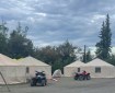 Image shows four tan round yurts that are being used for the incident command post of the Lush Fire. Stacks of supplies are under plastic tarps to the right and two four wheeler vehicles are in front of the yurts. The sky above is filled with grey clouds.