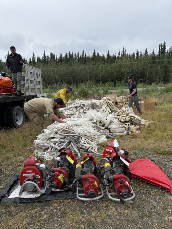 Five water pumps with a pile of hose in the background. Firefighters loading all onto a truck.