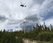 A helicopter hovers above a forested area with a water bucket dangling from a long line, while a firefighter observes from below.