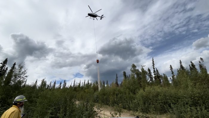 A helicopter hovers above a forested area with a water bucket dangling from a long line, while a firefighter observes from below.