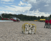 The image shows a cargo net filled with fire hoses, intended for use on the Ninetyeight Fire. In the background to the right, three firefighters are visible. To the left, a helicopter can be seen. The scene is set on a rocky creek bed.