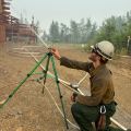 Firefighter wearing a white helmet, yellow shirt, and green pants sets up a sprinkler pointed at a log house