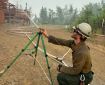 Firefighter wearing a white helmet, yellow shirt, and green pants sets up a sprinkler pointed at a log house
