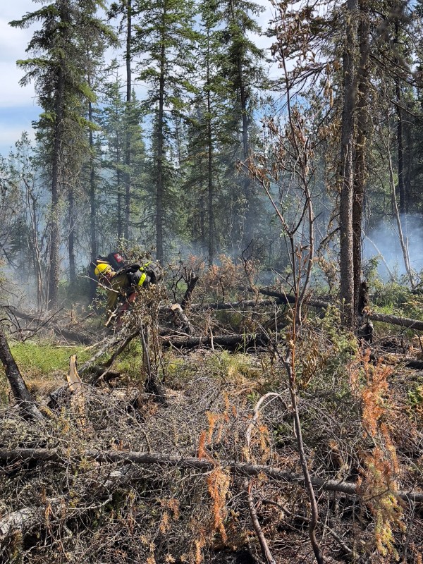 A firefighter in protective clothing works in a large brush pile with smoke in the background