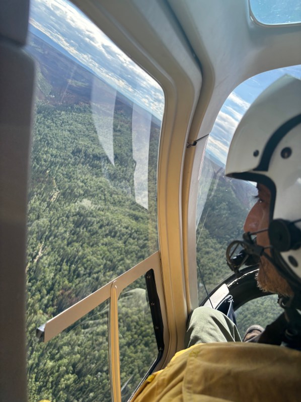 A firefighter looks out a helicopter window onto a forested and partially burned area below.