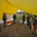Eight firefighters stand in a semicircle under a yellow tarp next to an information board with maps on it