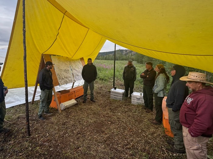Eight firefighters stand in a semicircle under a yellow tarp next to an information board with maps on it