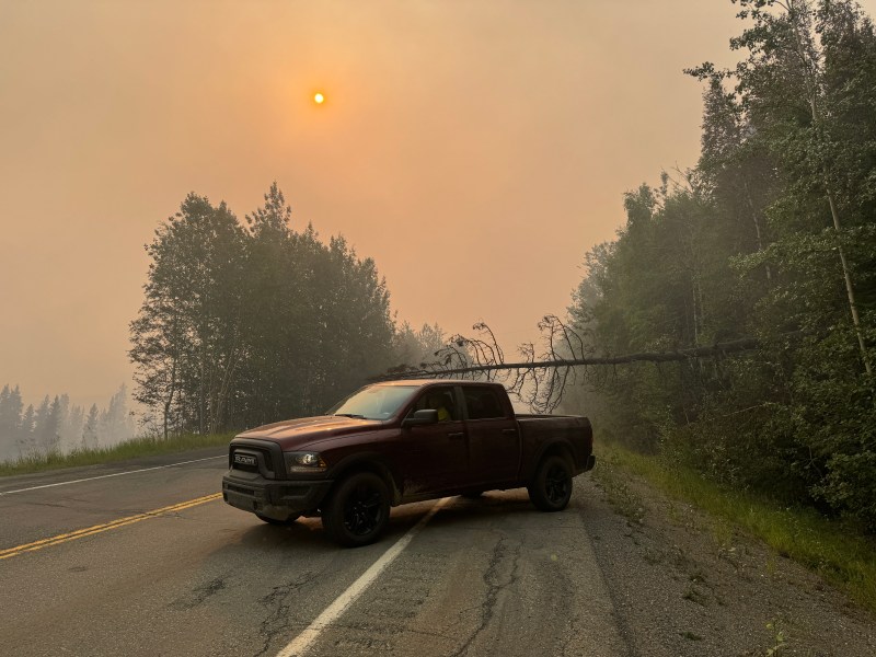 A truck is stopped as a fallen tree blocks the road. Heavy smoke cast an orange glow in the background.