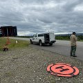 A man with a remote control in hand stands by the side of the road. Behind him is a landing zone for an Unmanned Aerial System.