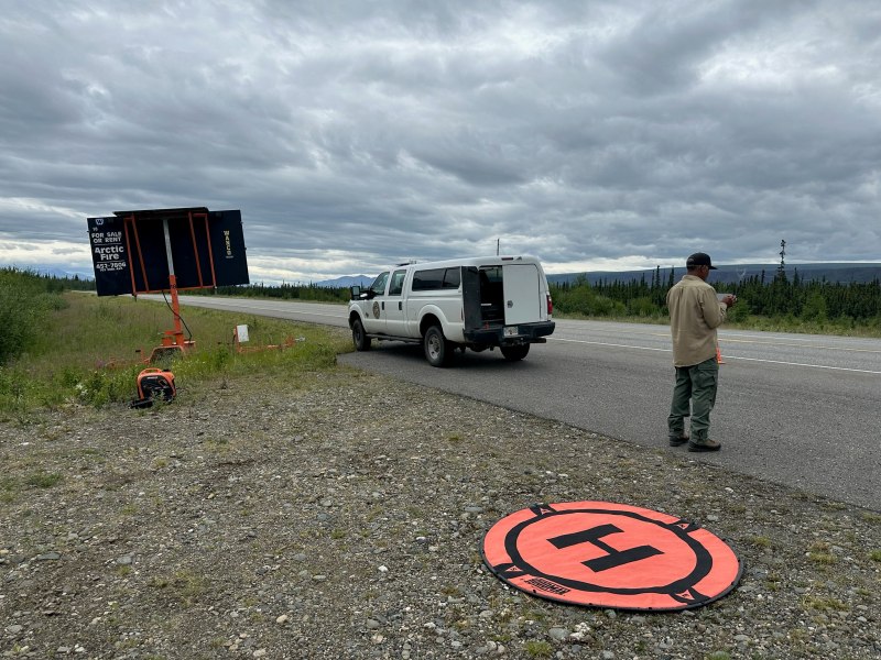 a man with a remote control in hand stands beside a road which contains a truck and a traffic message sign. Behind him is an orange plastic target with the letter h on it made of plastic sitting on rocks.